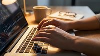 Professional programmer coding on laptop with warm lighting, hands typing on keyboard, coffee cup and glasses on desk creating productive workspace atmosphere.