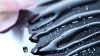 Glossy black liquid dripping in elegant curves with water droplets, creating smooth flowing streams against soft pink background for cosmetic design.