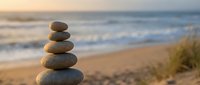 Balanced stone cairn on sandy beach at sunset with ocean waves and grass creating peaceful zen meditation atmosphere for wellness and mindfulness practices.