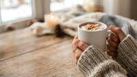 Person holding warm mug of coffee with latte art in cozy home interior with natural window light and soft furnishings.