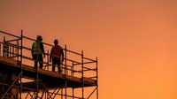 Construction workers silhouettes on scaffolding against dramatic orange sunset sky. Industrial building site with safety equipment and structural framework.