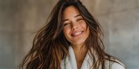 Young Hispanic woman with long wavy brown hair smiling joyfully in white bathrobe, expressing happiness and relaxation in spa or wellness setting.