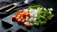 Fresh diced vegetables including orange carrots, white onions and green celery arranged on dark cutting board for cooking preparation and meal planning.