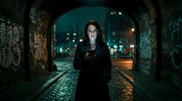 Young woman with long dark hair, South Asian ethnicity, wearing black jacket, standing in urban underpass at night holding smartphone, surrounded by graffiti walls and city lights in background, moody atmospheric street scene.