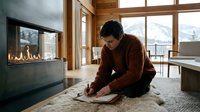 Young Asian man in cozy brown sweater reading book by modern fireplace in luxury mountain cabin with wooden interior and large windows.