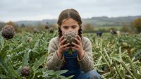 Young girl drinking hot beverage in autumn farm field with artichokes, cozy seasonal outdoor moment in countryside agricultural setting.