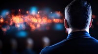 Back view of man looking at blurred city lights at night, dark silhouette against glowing red and blue bokeh background creating atmospheric urban scene.