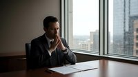 Thoughtful businessman in suit contemplating strategy while sitting at conference table near office window with city view during daytime meeting.