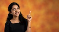 Young Indian woman in black top pointing upward with finger against warm orange background, smiling confidently for business presentation concepts.