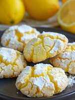 Homemade lemon crinkle cookies dusted with powdered sugar on dark plate with fresh lemons in background, showing distinctive crackled surface texture.