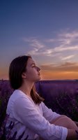 Young woman with long brown hair and Caucasian ethnicity wearing white shirt standing in lavender field at sunset, gazing upward at dramatic sky with purple and orange clouds during golden hour.