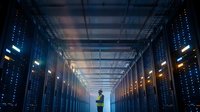 Male technician in safety vest working in modern data center server room with blue LED lighting and network infrastructure for cloud computing services.