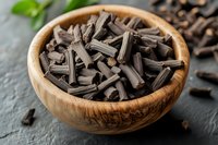 Dried licorice root pieces in wooden bowl on dark stone background with fresh mint leaves, natural herbal medicine and traditional spice ingredient.