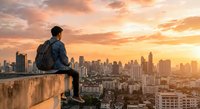 Young Asian man with backpack sitting on rooftop edge overlooking urban cityscape at golden hour sunset. Contemplative traveler enjoying panoramic city view.