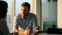 Professional businessman in casual shirt having focused conversation with colleague in modern office setting with natural window lighting.