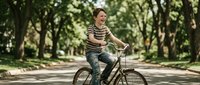 Happy young Caucasian man in striped shirt riding vintage bicycle through tree-lined park path on sunny day, enjoying outdoor recreation and healthy lifestyle.