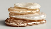 Stack of golden and pearl liquid lipgloss swatches on neutral background, showcasing different metallic and shimmer finishes for makeup artistry and beauty product photography.