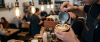 Barista pouring latte art into coffee cup at busy cafe counter with warm Edison bulb lighting and blurred customers in background.