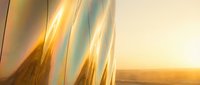 Iridescent metallic surface reflecting golden sunset light, futuristic architecture detail with shimmering chrome finish against warm desert horizon.