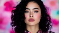 Young Asian woman with curly black hair and natural makeup against pink blurred bokeh background, looking directly at camera with confident expression.
