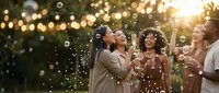 Group of diverse young adults celebrating outdoors with champagne glasses and soap bubbles at sunset party with warm bokeh lights in background.