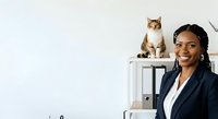 Young Black female professional in dark blazer and white shirt smiling at camera in modern office with tabby and white cat sitting on shelf behind her.