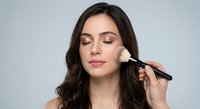 Young brunette woman applying makeup with brush on face, eyes closed, professional beauty routine for cosmetics and skincare advertising.