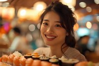 Young Asian woman smiling at restaurant with sushi platter in foreground, warm ambient lighting creates festive dining atmosphere.