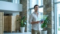 Young professional businessman holding tablet computer in modern office with stone walls and large windows during daytime work session.