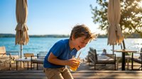 Young Caucasian boy in blue shirt enjoying orange juice at waterfront restaurant terrace during golden hour sunset with lake view and outdoor dining area.