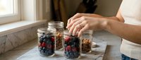 Woman preparing healthy breakfast parfait with fresh berries and granola in glass jars on kitchen counter for meal prep and nutritious eating lifestyle.