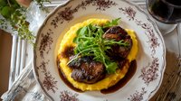 Seared foie gras on creamy polenta with arugula garnish and red wine reduction sauce, served on elegant porcelain plate with floral pattern.