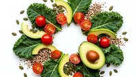 Fresh healthy food ingredients arranged in circle with avocado slices, cherry tomatoes, kale leaves and seeds on white background for nutrition concepts.