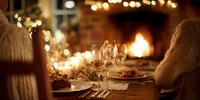 Elegant dinner table with wine glasses by warm fireplace, festive string lights creating cozy holiday atmosphere for intimate gathering.