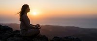 Young woman meditating in lotus pose on mountain rocks during golden sunset, practicing mindfulness and yoga outdoors for wellness and spiritual peace.