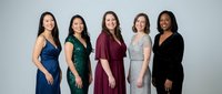 Five diverse professional women in elegant formal dresses standing together against neutral background, representing teamwork, success and corporate diversity.