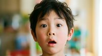 Young Asian boy with messy hair showing surprised facial expression with wide eyes and open mouth in bright indoor setting for emotional reactions.