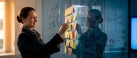 Professional businesswoman organizing colorful sticky notes on glass wall during strategic planning session in modern office environment.