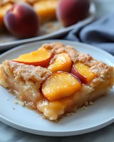 Homemade peach upside-down cake slice topped with caramelized fruit and cinnamon sugar, served on white plate with fresh peaches and napkin in background.