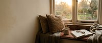 Cozy reading nook by window with warm sunlight, soft pillows and open book creating peaceful home interior atmosphere for relaxation and leisure time.