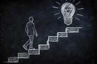 Businessman climbing stairs towards glowing lightbulb drawn on blackboard, representing career growth, innovation and path to success in business concept.