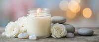 Spa wellness scene with burning candle in glass jar, white roses, smooth stones and soft bokeh lights creating peaceful relaxation atmosphere for meditation.