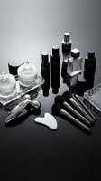 Luxury skincare and cosmetic products collection with black bottles, glass containers, and beauty tools arranged on reflective surface with gradient background.