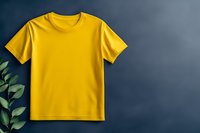 Yellow cotton t-shirt mockup on dark background with green plant leaves, blank apparel template for branding and design presentation.