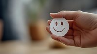 Female hand holding white paper smiley face emoji against blurred background. Positive mood and happiness concept for mental health and wellbeing content.