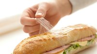 Caucasian hand sprinkling water or seasoning onto fresh baguette sandwich with ham and lettuce, food preparation and cooking concept.