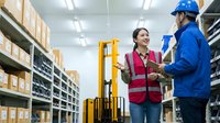 Asian female warehouse manager in safety vest discussing logistics with male worker in blue uniform and hard hat in modern distribution center.