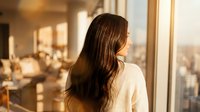 Young woman with long brown hair in white sweater looking out office window at sunset cityscape with warm golden light streaming through glass.