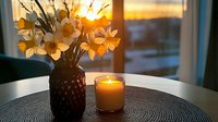 Cozy home interior with spring flowers in vase and glowing candle on table during golden sunset through window creating warm atmospheric lighting.