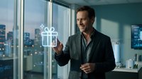 Middle-aged man with dark hair and facial stubble, wearing a dark blazer over a black shirt, gesturing toward a glowing gift box icon in a modern high-rise office with city skyline views. Suitable for business, technology, rewards, and corporate concepts.